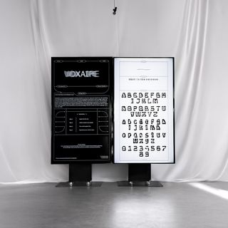 Installation von »VOXAIPE – Decoded« mit zwei Monitoren; Foto: Mai Do Zwei nebeneinander stehende Monitore vor weißem Stoffhintergrund. Links ein dunkles Interface von VOXAIPE, rechts eine helle Seite mit generierten Buchstabenformen.
