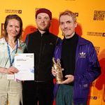 Laura Bein (Schauspielerin), Valentin Bolte (Regie und Kamera) und Georg Kästle (Regie und Drehbuch) auf der Preisverleihung in Dresden. Bild: Elena Pagel Zwei Männer und eine Frau stehen vor einer gelben Fotowand beim Filmfest Dresden. Die Frau hält eine Urkunde, einer der Männer eine Trophäe.