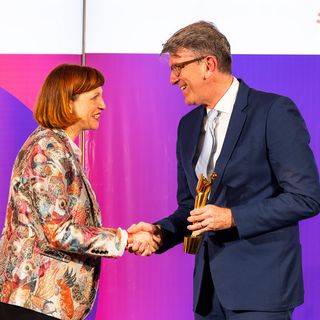 Der Staatsminister für Kultur und Medien, Wolfram Weimer, überreichte den Preis an Alumna Lena Libertà. Foto: BKM /Chiara Lang Lena Libertà schüttelt Kulturstaatsminister Wolfram Weimer auf einer Bühne die Hand. Wolfram Weimer hält eine goldene Preisfigur, beide lächeln vor einer farbigen Hintergrundwand.
