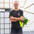 Jakob Elias Meyer ist für seine Arbeit »Subsistenzprovisorium« mit den Lyonel Kunstpreis 2026 ausgezeichnet worden. Foto: Matthias Eckert Jakob Elias Meyer steht mit verschränkten Armen vor einer Installation aus Holzlatten und einem großen Kunststoffbehälter. In einer Hand hält er einen Blumenstrauß und eine leuchtend gelbe Urkunde.