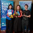 v.l.n.r. Tara Thomson, Anya Ryzhkova und Felix Weller auf der Preisverleihung des Kurzsuechtig-Festivals. Foto: Anika Dollmeyer / Kurzsuechtig Tara Thomson, Anya Ryzhkova und Felix Weller stehen bei der Preisverleihung des Kurzsuechtig-Festivals vor Veranstaltungsbannern, halten Urkunden, Blumen und einen Preis in den Händen und blicken in die Kamera.