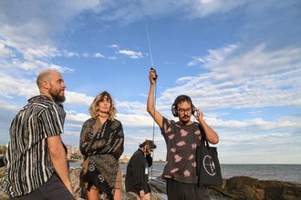 Vier Menschen stehen am Meer, einer hält ein tragbares Radio mit ausgefahrener Antenne in die Höhe und lauscht über Kopfhöhrer, die er trägt