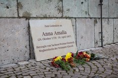 Larissa Barth's commemorative plaque for Anna Amalia leans against the outer wall of the Anna Amalia Library. On the cobblestone pavement in front are red and yellow flowers. The inscription on the lime sandstone plaque reads: In memory of Anna Amalia (24 October 1739 - 10 April 1807) / Duchess of Saxony.Weimar-Eisenach / Pioneer of Weimar Classicism.
