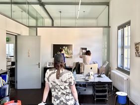 The »nova space« curatorial team sets up the exhibition. Photo: Benedict Thiele Two people with white cotton gloves align two framed photographs on a wall, another person at a distance provides information as to whether the pictures are hanging straight