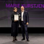 Foto: Salar Baygan Marie Kurstjens steht bei der Preisverleihung des German Design Award 2026 auf der Bühne und hält ihre Urkunde sowie die Trophäe als »Newcomer of the Year«; neben ihr steht ein Vertreter der Veranstaltung, im Hintergrund ist ihr Name groß projiziert.