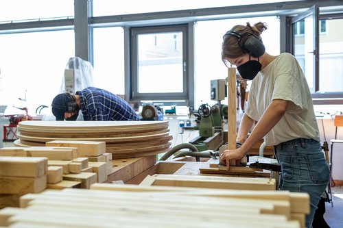 Eine Studentin arbeitet mit Mundschutz in der Holzwerkstatt an einem Möbelstück für die das Foyer der Mensa, im Hintergrund ist ein Student zu sehen. Bauhaus-Universität Weimar, Foto: Thomas Müller