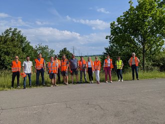 Frauen und Männer stehen am Rand einer Straße. Manche von ihnen tragen Kleidung in Signalfarbe.