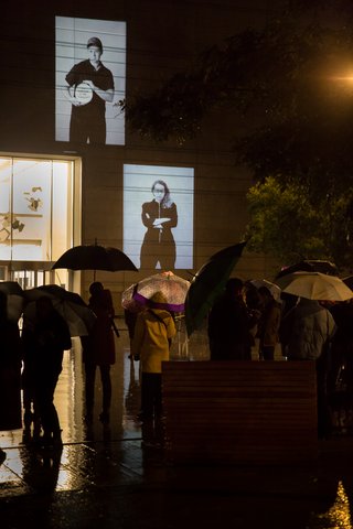 The photo shows 2 black-and-white portraits from Larissa Barth's photo series walter ≠ bauhaus, which are projected onto the on the façade of the Bauhaus Museum Weimar at night. Passers-by with umbrellas are standing in the square outside the museum, looking at the pictures.