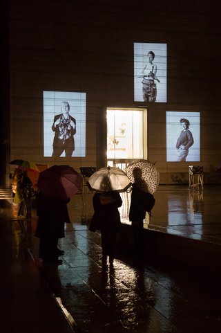 The photo shows 3 black-and-white portraits from Larissa Barth's photo series walter ≠ bauhaus, which are projected onto the on the façade of the Bauhaus Museum Weimar at night. Passers-by with umbrellas are standing in the square outside the museum, looking at the pictures.