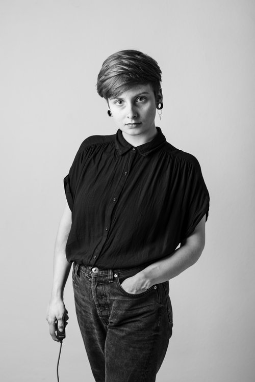 The black-and-white photograph shows concept artist Larissa Barth. Barth has short, dark-blond hair and black tunnel earrings. She is wearing dark jeans and a blouse made of a thick, black cloth with short sleeves. Her left hand is in the pocket of her jeans. Her right hand is holding the self-timer. She is looking self-confidently into the camera lens.
