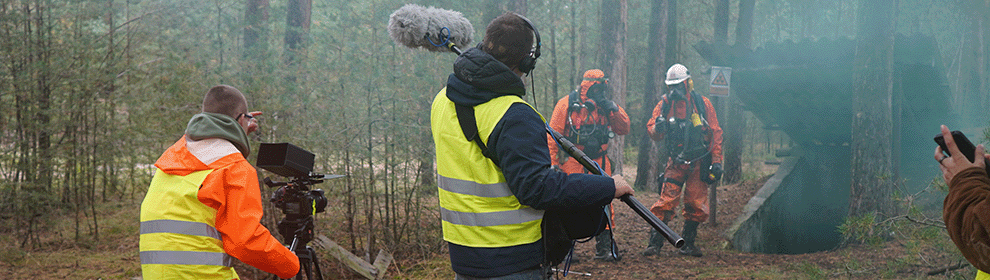 Zwei Studierende mit Kamera und Mikrofon stehen mit Warnwesten bekleidet im Wald und nehmen auf, wie zwei Männer in orangenen Anzügen und Atemschutzmasken aus einem Bunker laufen, aus dem dunkelgrüner Rauch kommt.